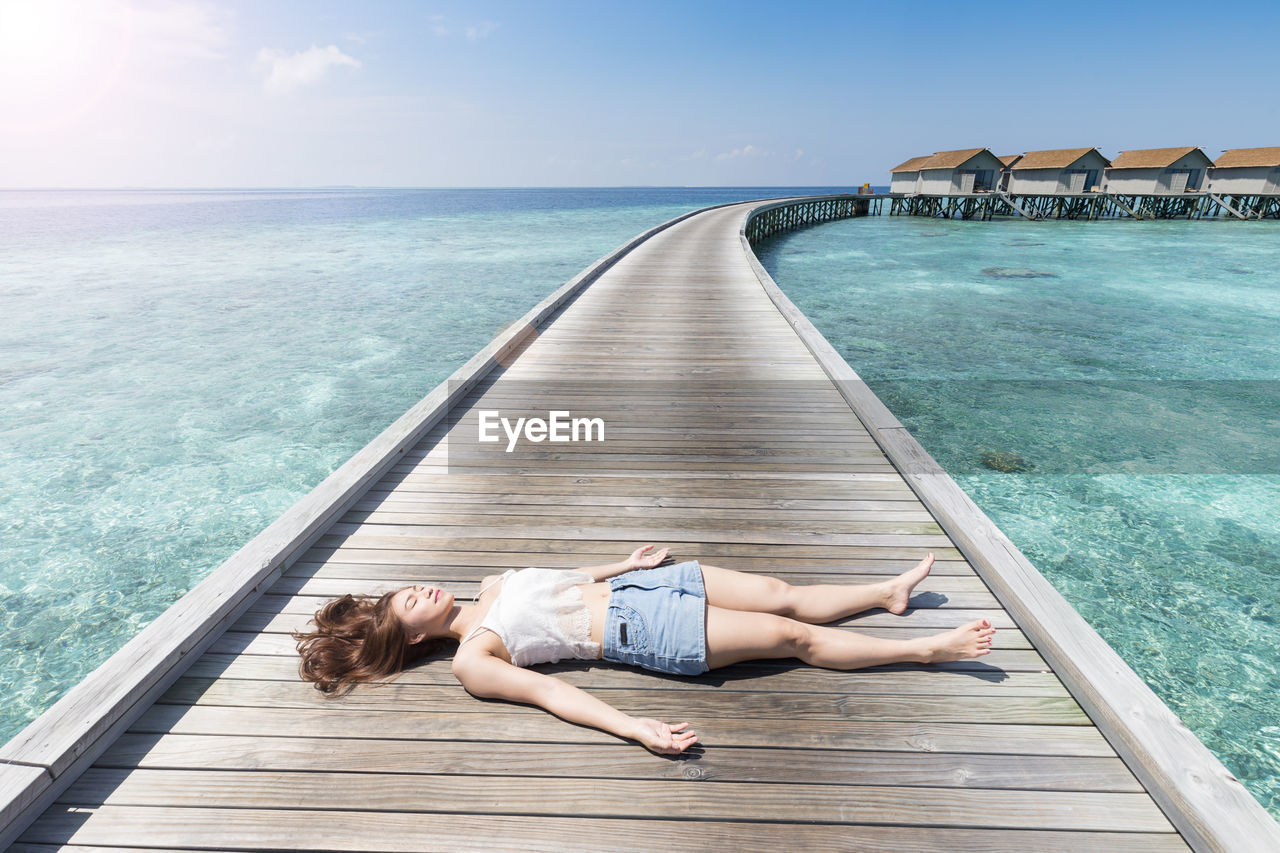 Woman lying on boardwalk amidst sea against sky