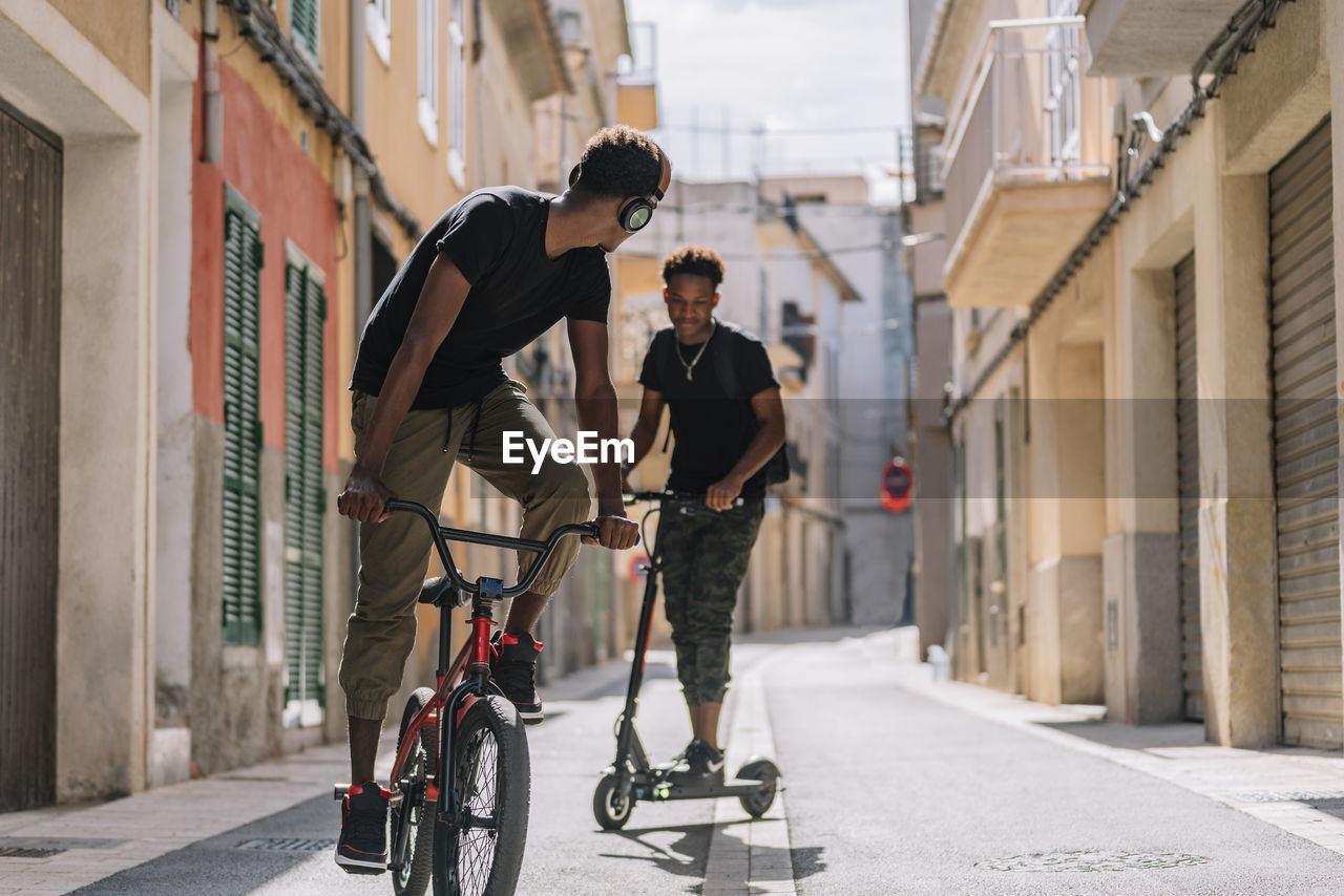 Cheerful young african american man riding electric scooter while black male friend is driving bicycle in street