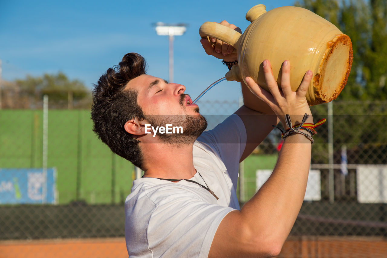 Young man drinking water from pot at court during sunny day