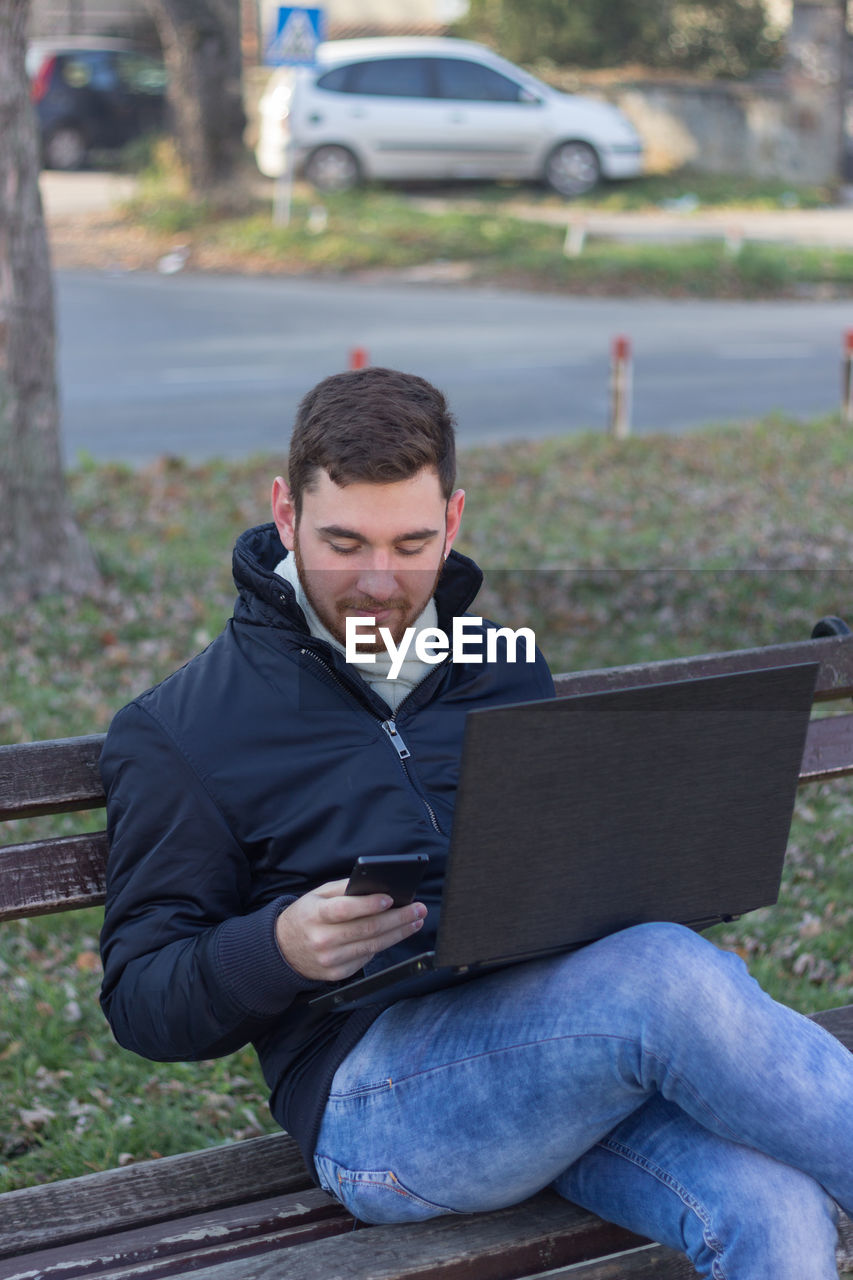Man with laptop using phone while relaxing on bench