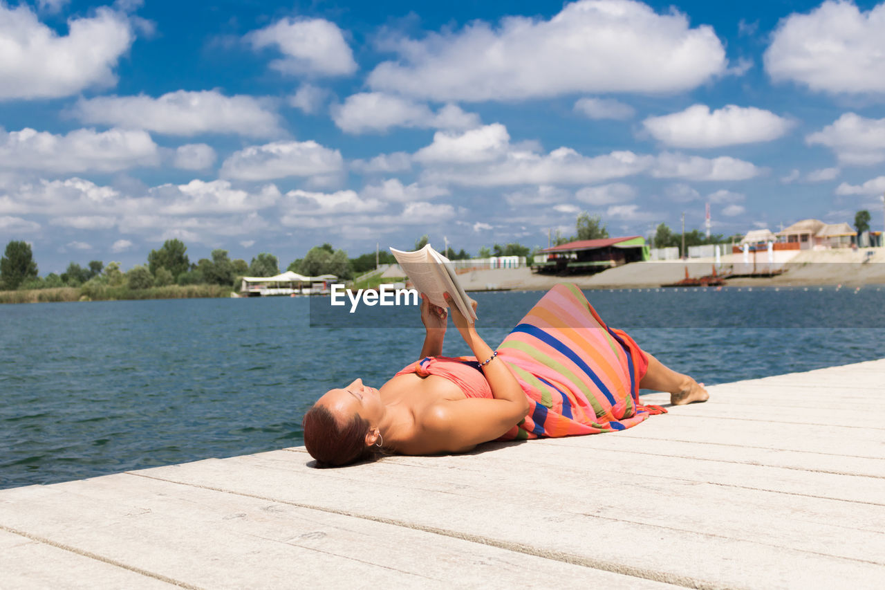 Woman reading book while lying on pier over sea against cloudy sky