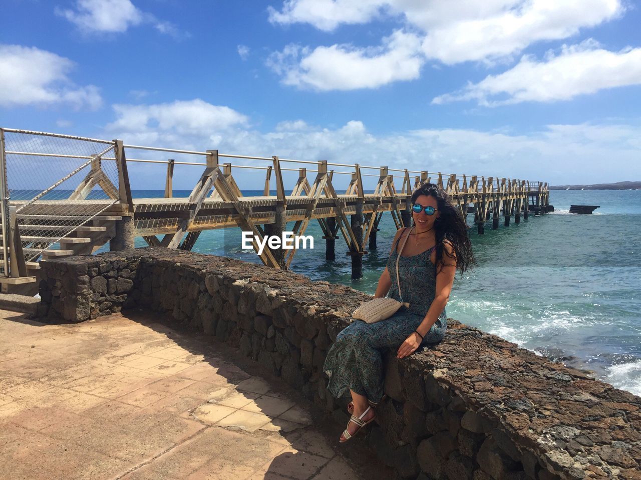 Portrait of beautiful young woman sitting on retaining wall against sea