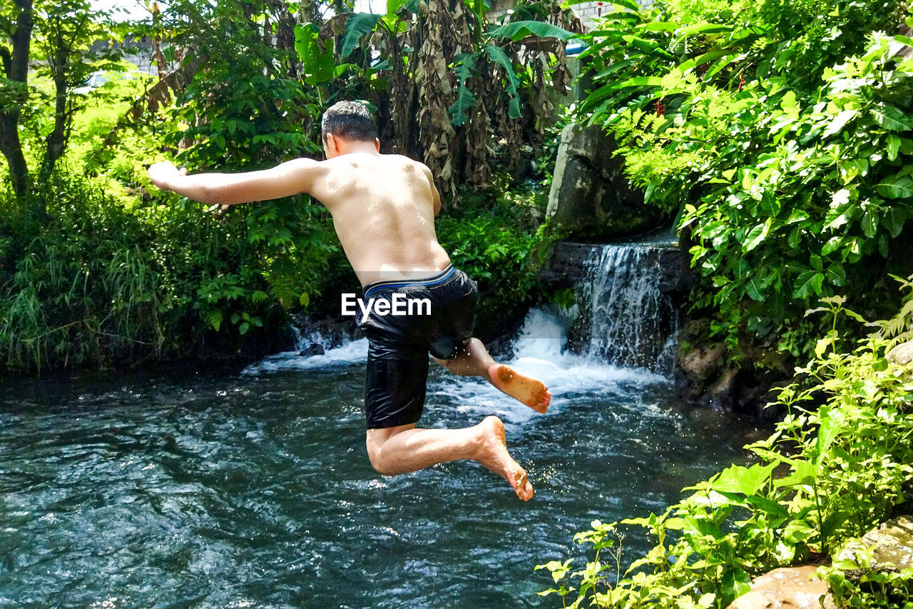 water, one person, nature, plant, tree, motion, leisure activity, men, jungle, full length, jumping, lifestyles, day, mid-air, forest, fun, waterfall, enjoyment, holiday, beauty in nature, vacation, outdoors, trip, young adult, adult, carefree, rainforest, swimming trunks, shorts, land, adventure, swimwear, river, vitality, green, sunlight, sports, splashing, casual clothing, summer, swimming, rock, growth, scenics - nature