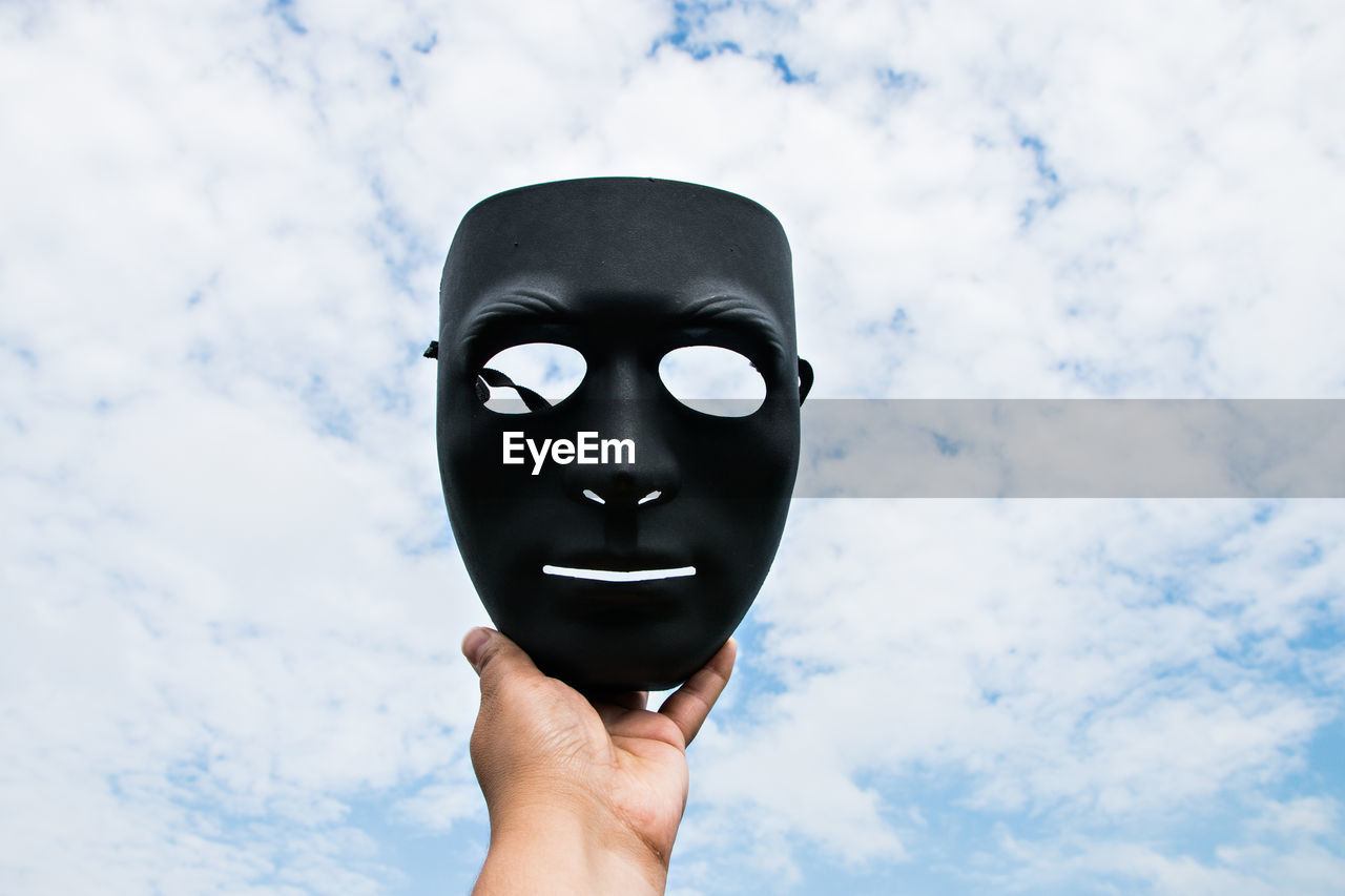 Cropped hand of person holding black mask against cloudy sky