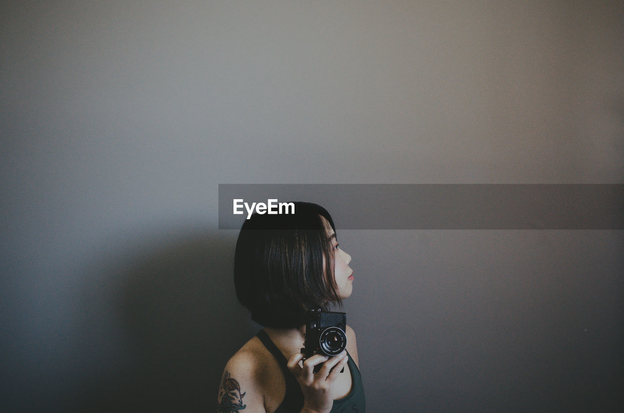 Woman holding camera while standing by wall