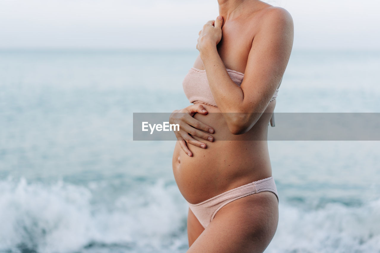 Close-up belly of a pregnant woman standing by the sea on the beach.