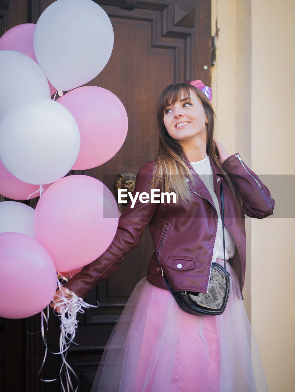 Smiling young woman holding pink balloons while standing against door
