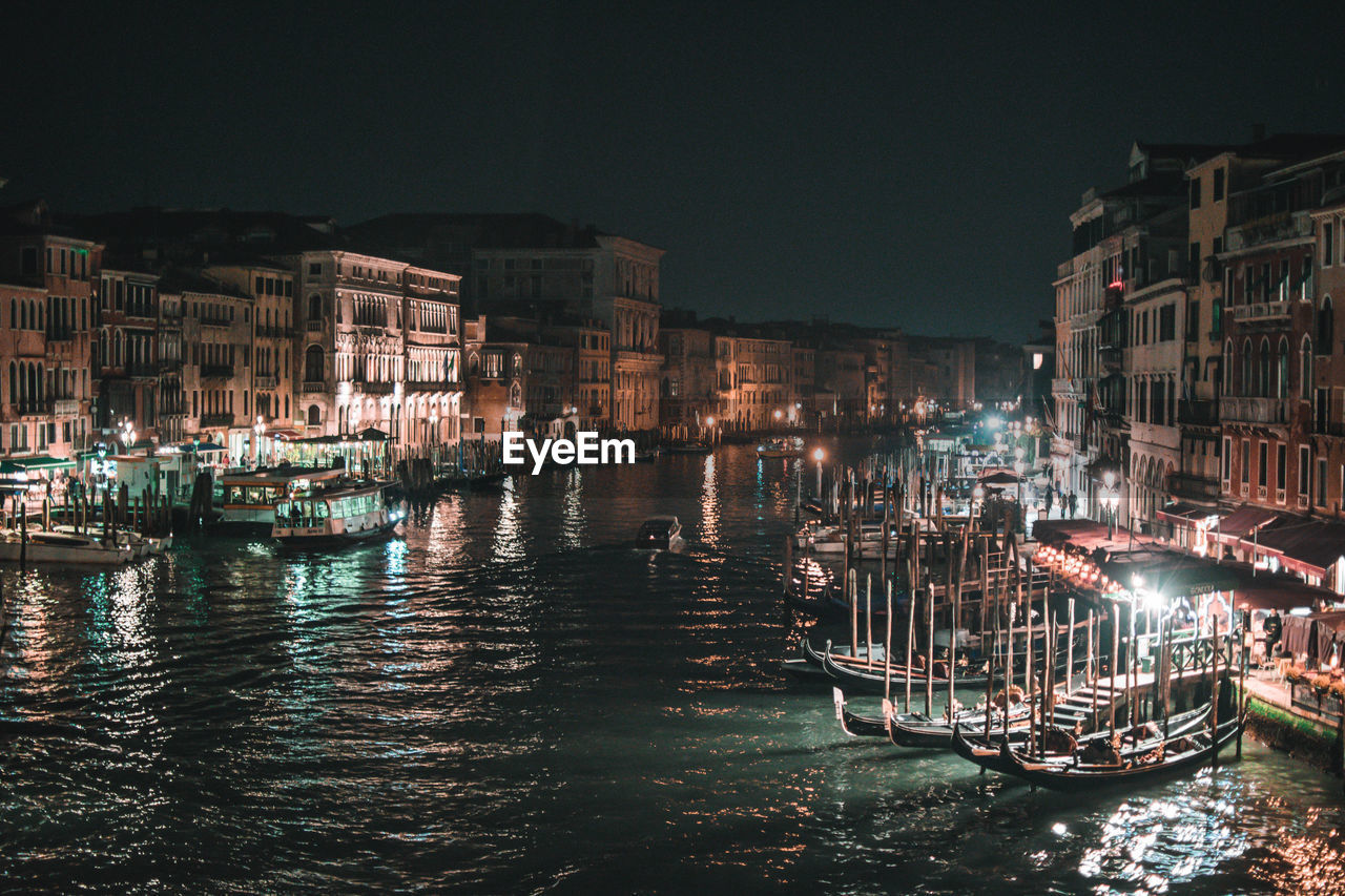 SAILBOATS MOORED ON CANAL BY ILLUMINATED CITY BUILDINGS AT NIGHT