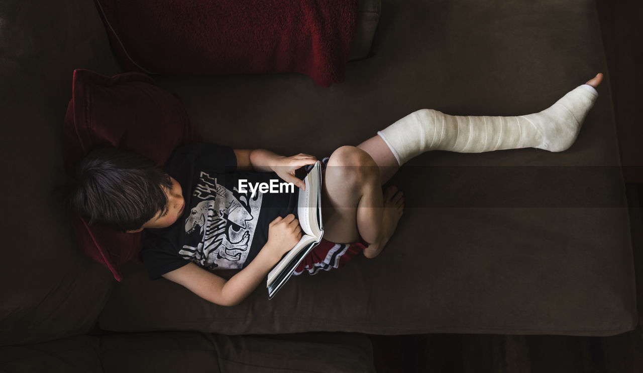 High angle view of boy with broken leg reading book while lying on sofa at home