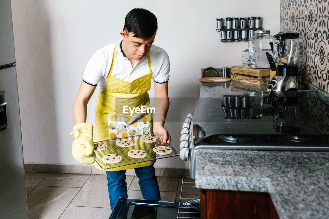 Ethnic teenage boy with down syndrome putting raw chocolate chip cookies in oven while baking pastry in kitchen