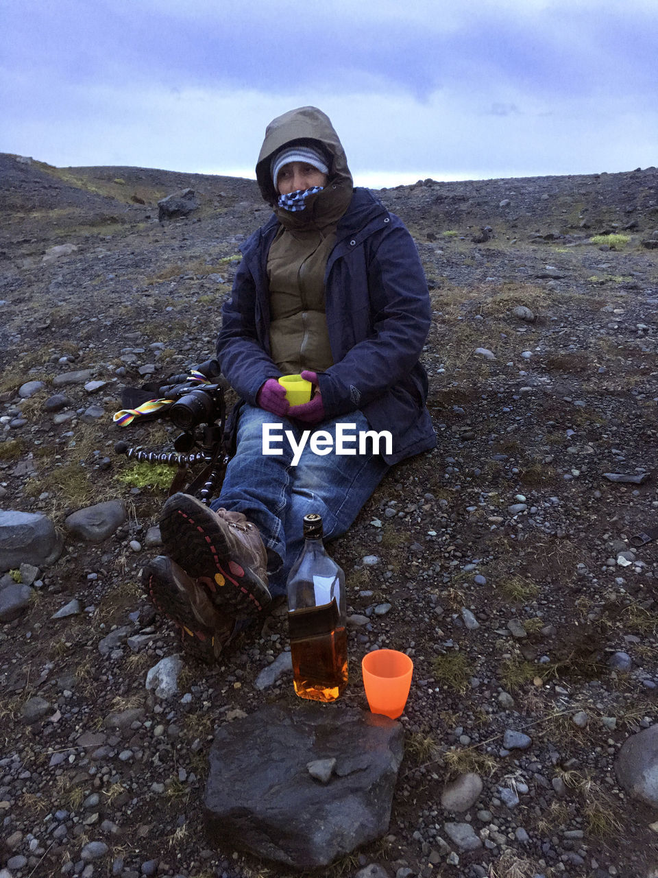 Full length of woman wearing warm clothing sitting on mountain with alcoholic drink
