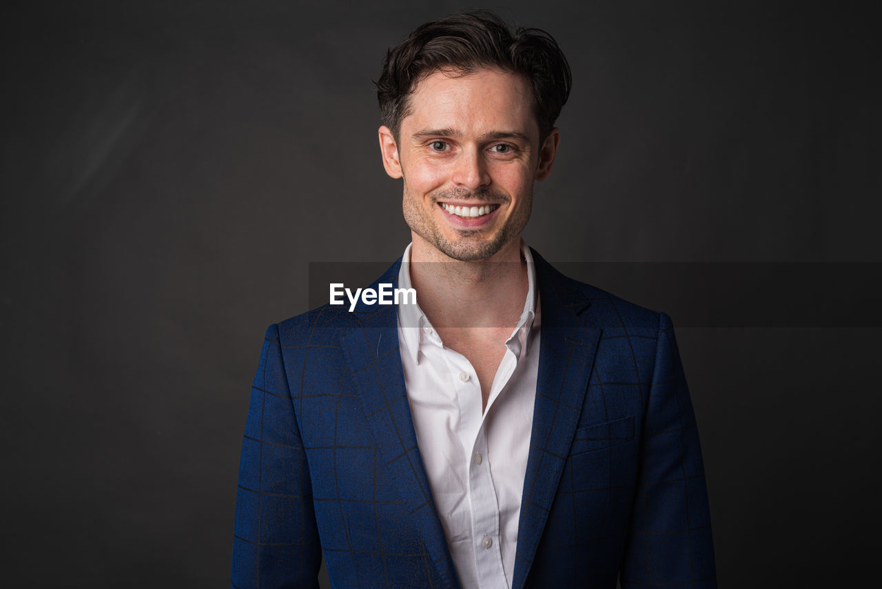 portrait, smiling, one person, adult, happiness, studio shot, looking at camera, business, men, indoors, formal wear, emotion, cheerful, businessman, front view, person, smile, teeth, gray, beard, button down shirt, white-collar worker, copy space, positive emotion, clothing, waist up, human face, success, headshot, standing, brown hair, business finance and industry, gray background, professional occupation, young adult, stubble, blazer, occupation, facial hair, black background