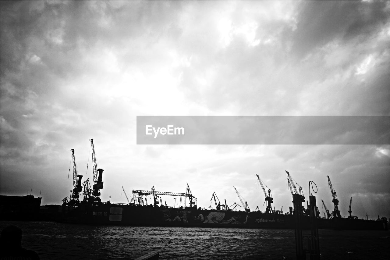 Cranes at commercial dock against cloudy sky