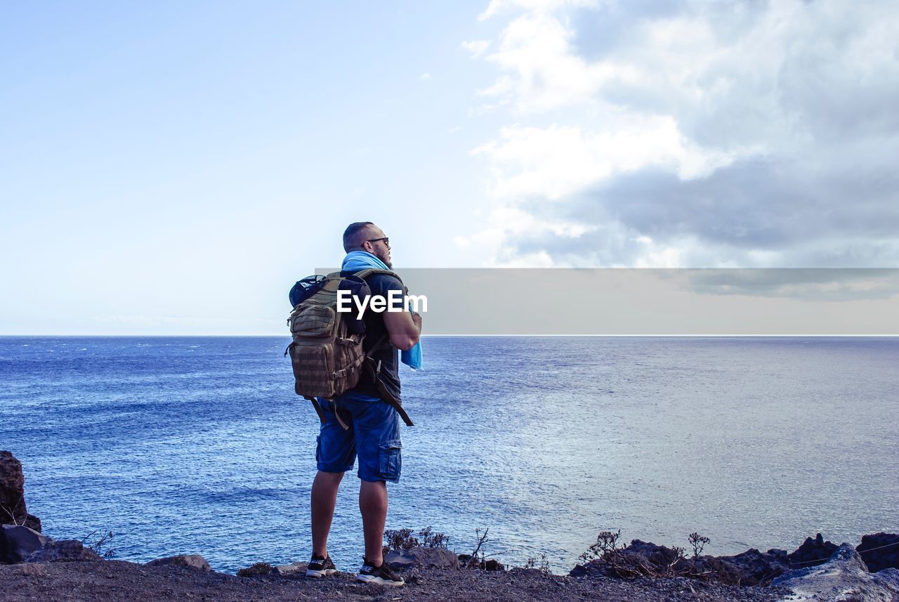 REAR VIEW OF MAN STANDING ON SHORE AGAINST SEA