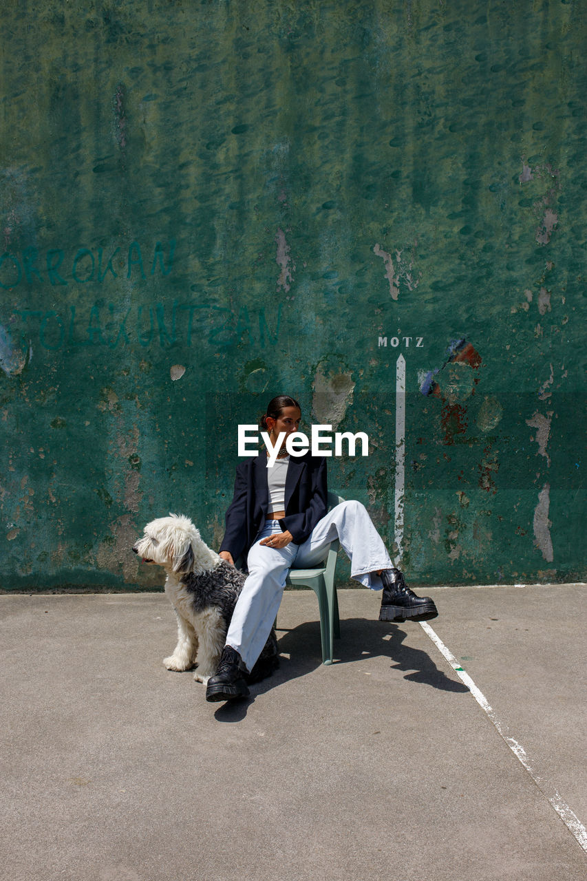 Full body of fashionable young ethnic female with dark hair in stylish outfit sitting on stool near adorable fluffy old english sheepdog near weathered wall on sunny day