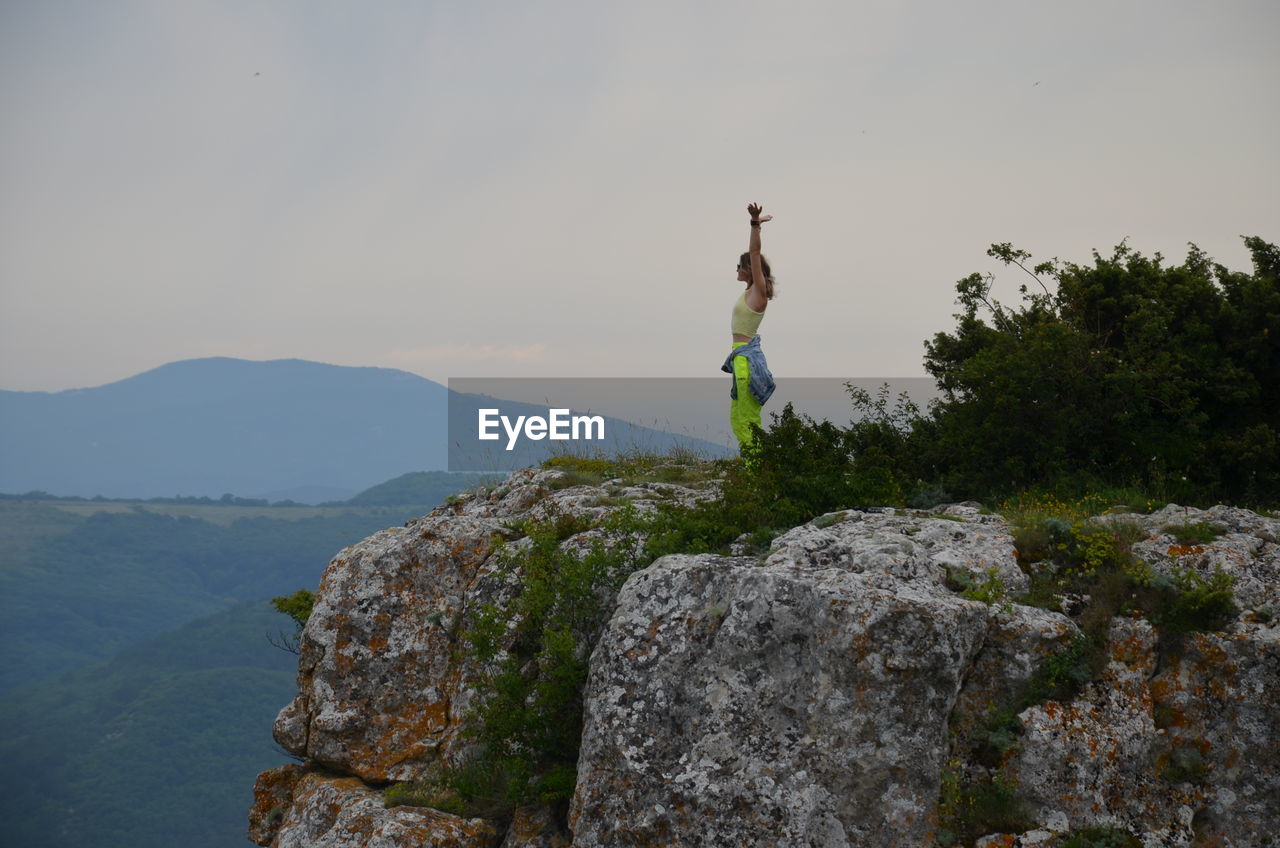 Wife jumping on rock by mountain against sky