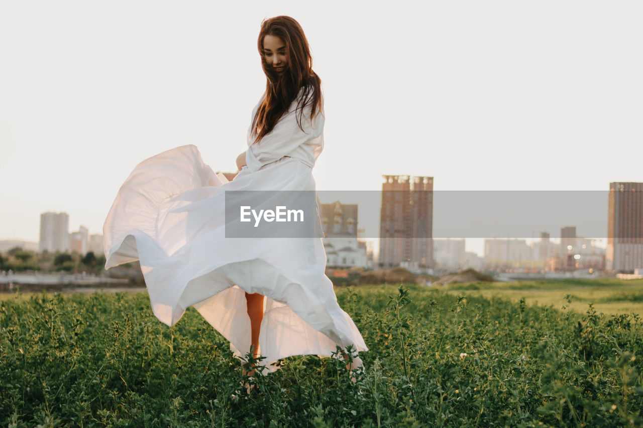 Full length of beautiful woman in white dress waking on land in city against clear sky during sunset