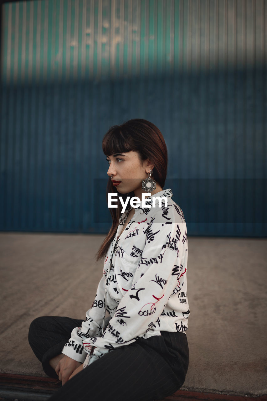 Dreamy young ethnic female millennial with long dark hair in casual clothes sitting on floor and looking away on street