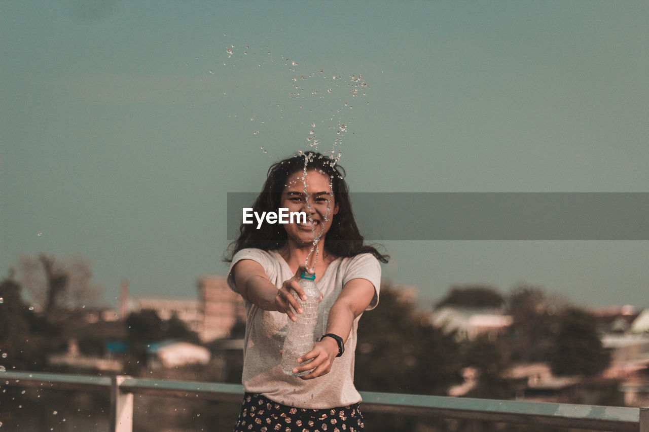 Portrait of smiling young woman throwing water against clear sky