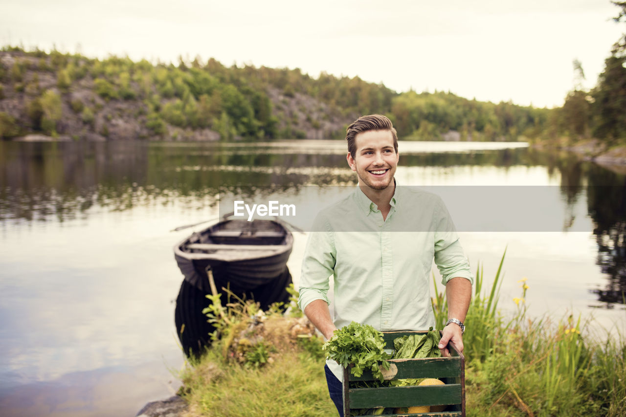 Smiling man carrying vegetables crate while standing against boat at lake