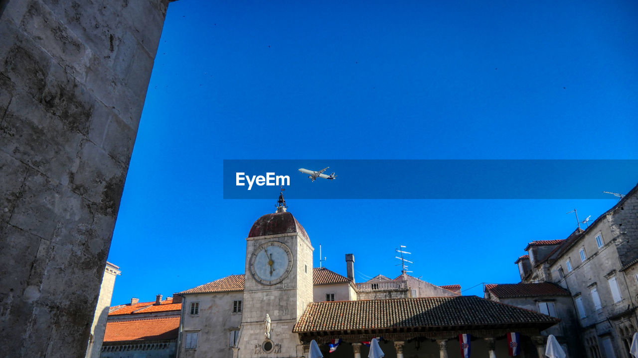 Low angle view of aeroplane flying by buildings against clear blue sky in trogir