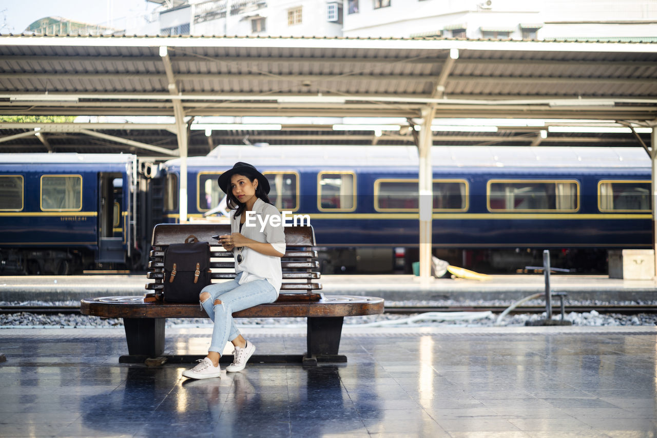Woman in white shirt sitting on wooden bench holding smartphone and thinking about her destination.