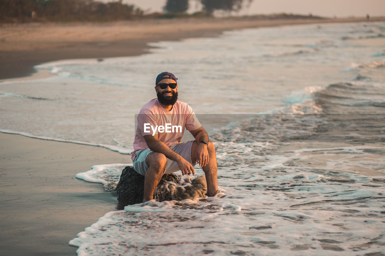 Full length of good looking bearded man sitting at the beach during sunset with feet in the water