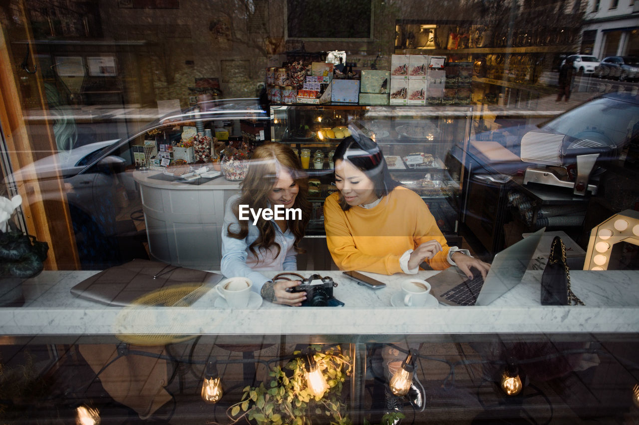 Female bloggers with camera and laptop sitting at cafe seen through window glass