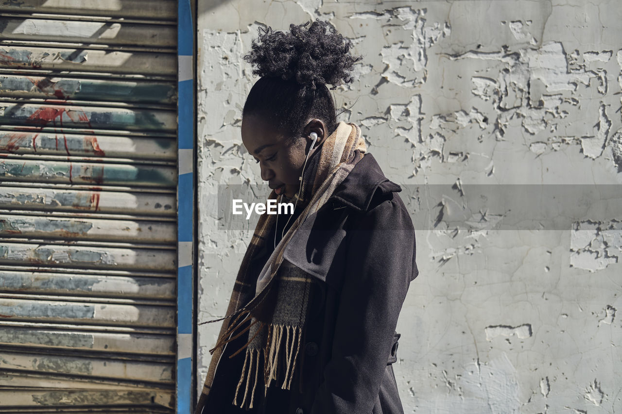 Side view of young attentive ethnic female in scarf with afro hair bun listening to song in earphones in town while looking down