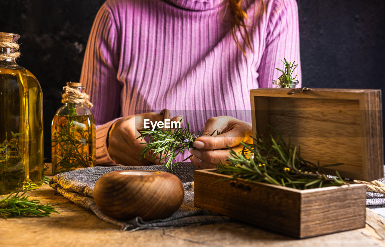 Crop lady cutting rosemary sprigs with scissors at table with fabric and rope near essential oil glass bottles and small chest