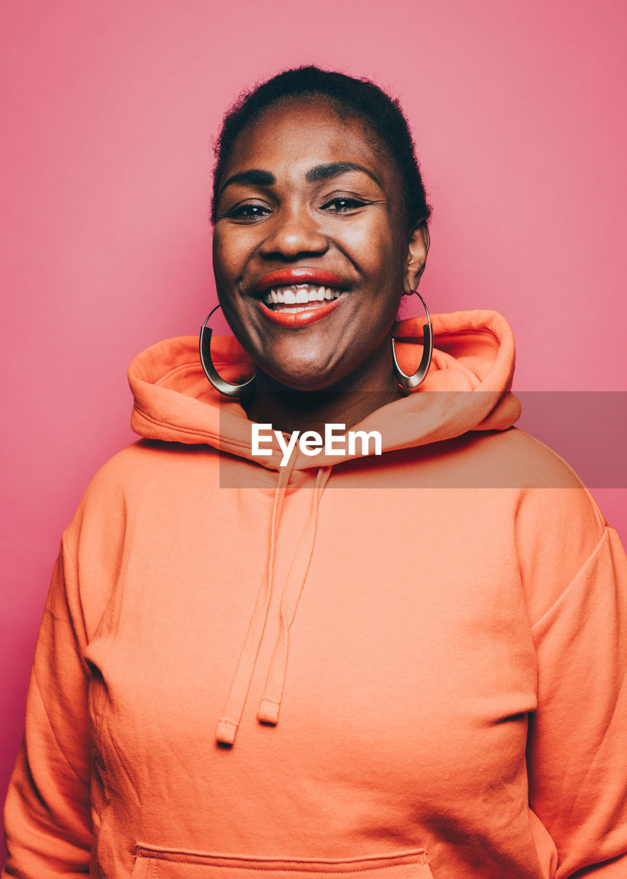 Portrait of smiling mid adult woman wearing orange hooded jacket over pink background