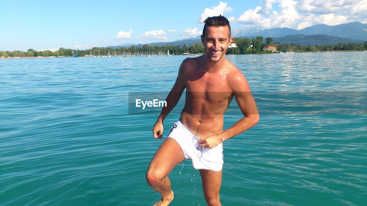 Portrait of smiling shirtless man in lake against sky