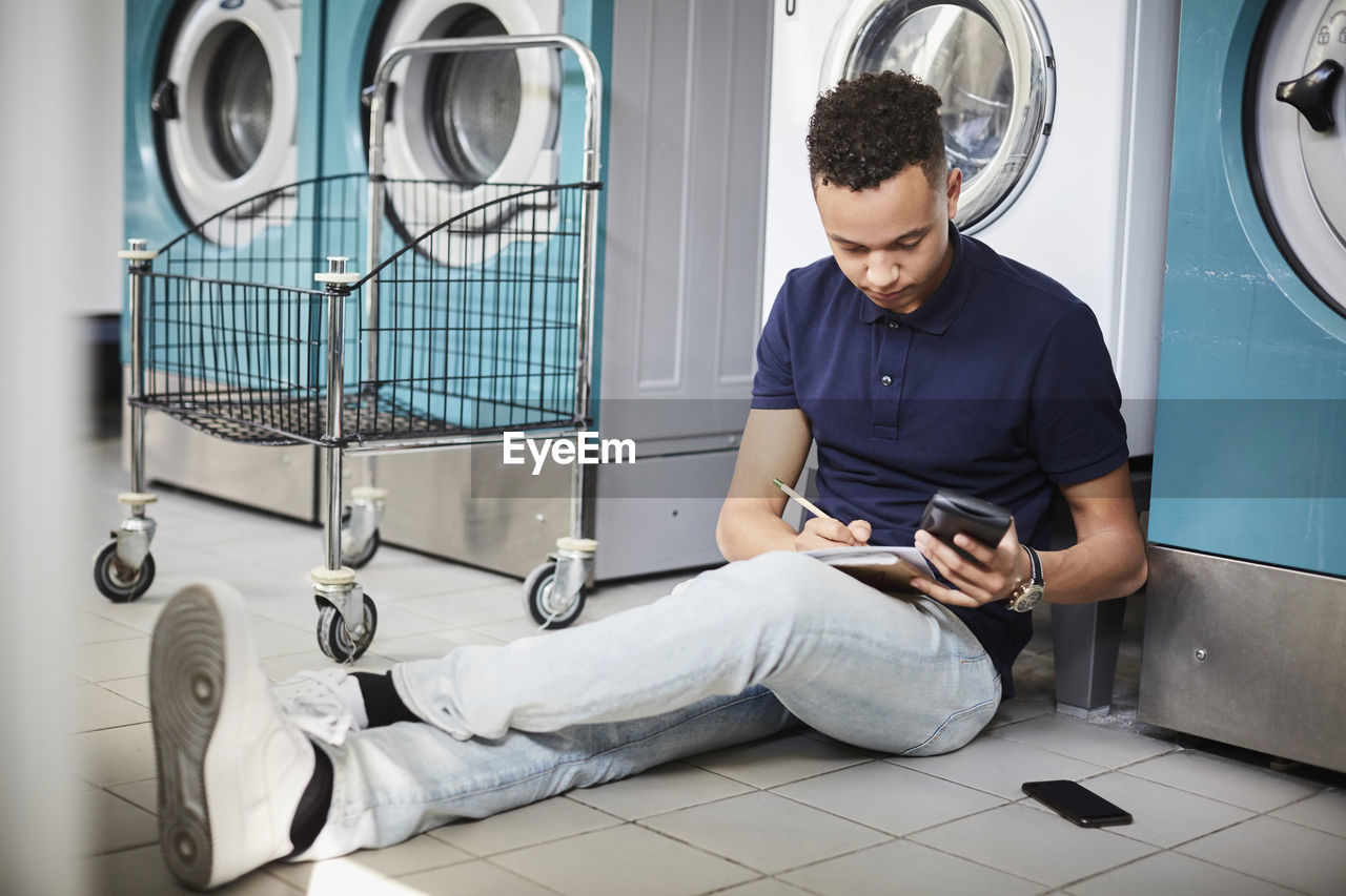 Young man holding mobile phone and studying while sitting at laundromat