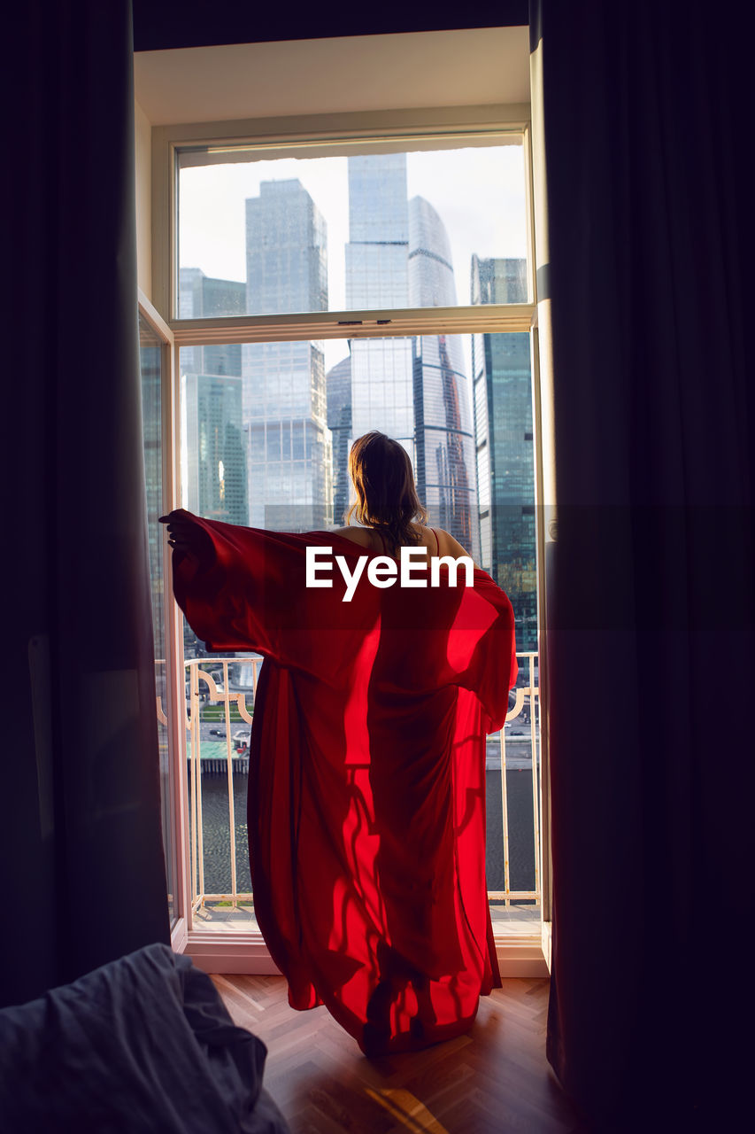 Brunette woman in a red bathrobe stands at a large window in an apartment opposite a skyscraper