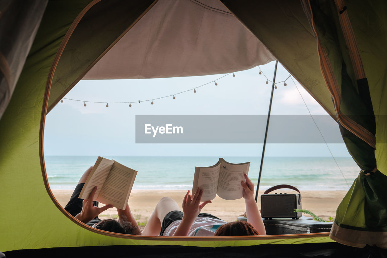 Asian couple camping and reading book on beach with sea background on summer vacation