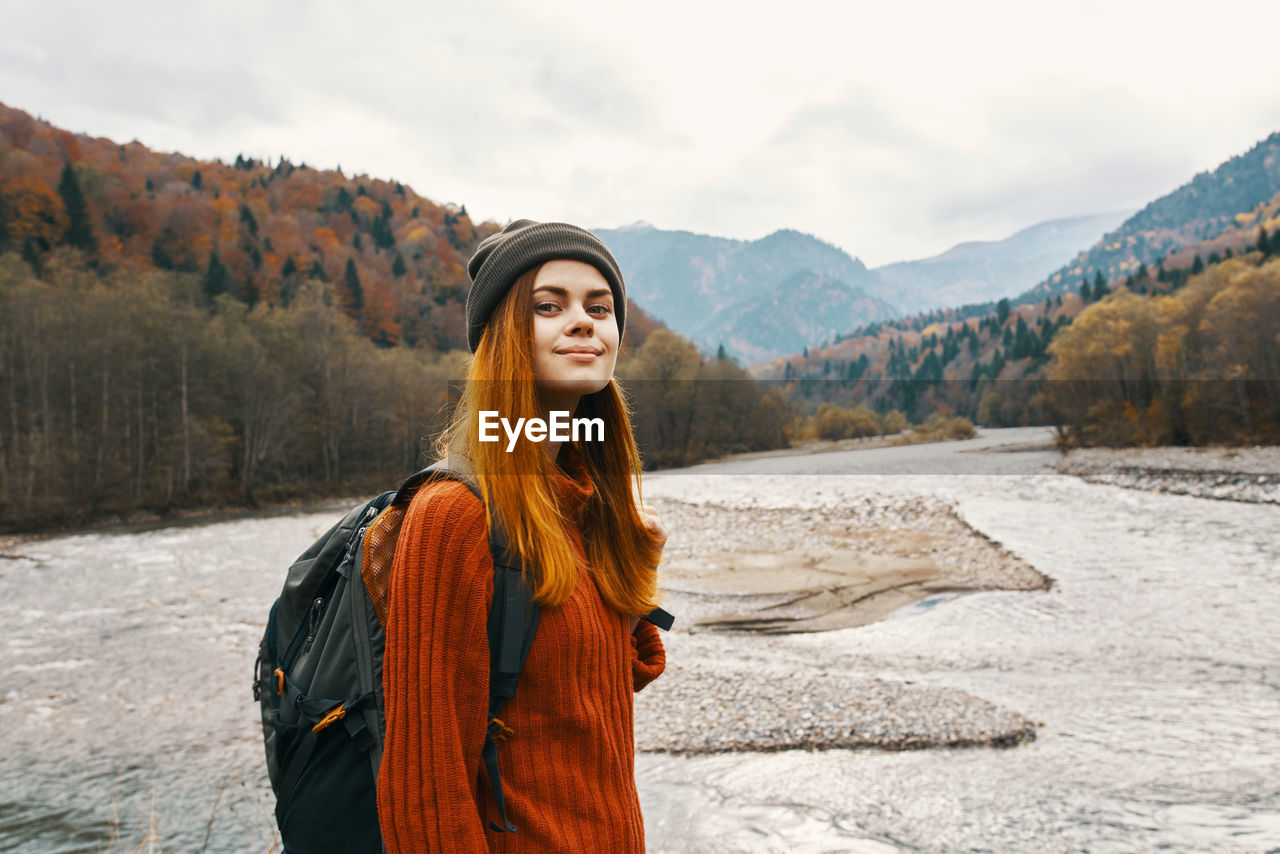 PORTRAIT OF SMILING YOUNG WOMAN STANDING AGAINST MOUNTAIN