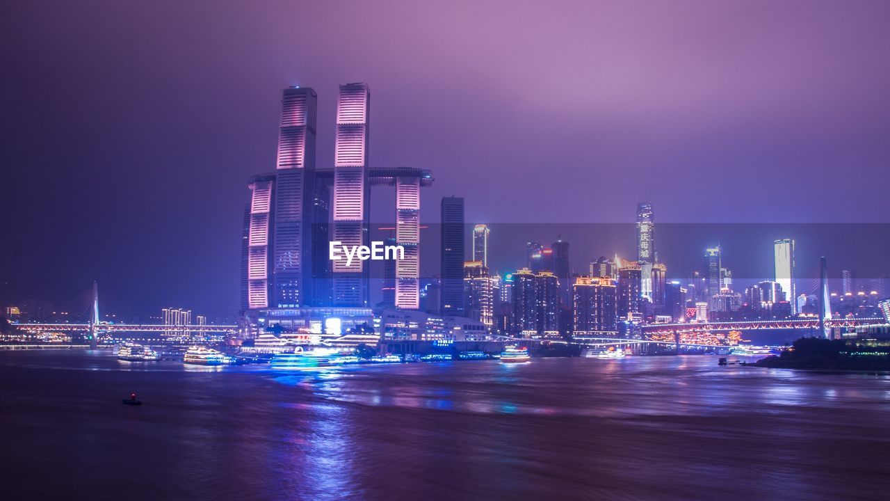 Illuminated modern buildings by bay against sky at night