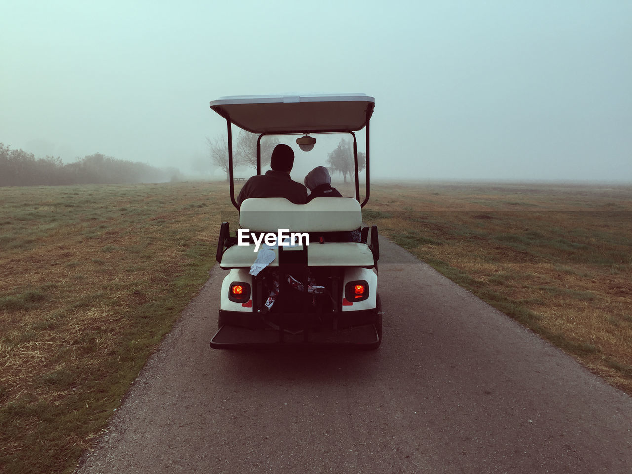 Rear view of father with son in golf cart on road amidst field during foggy weather