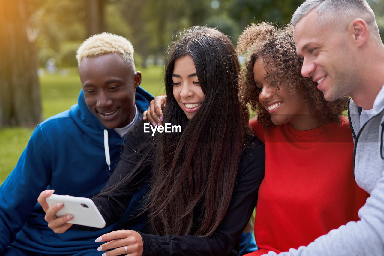 Smiling friends looking at phone sitting on bench at park