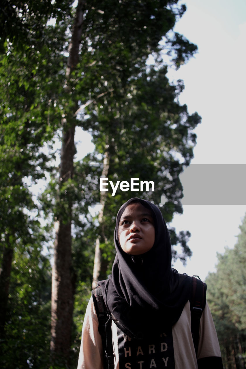 Low angle view of young woman in hijab standing against trees