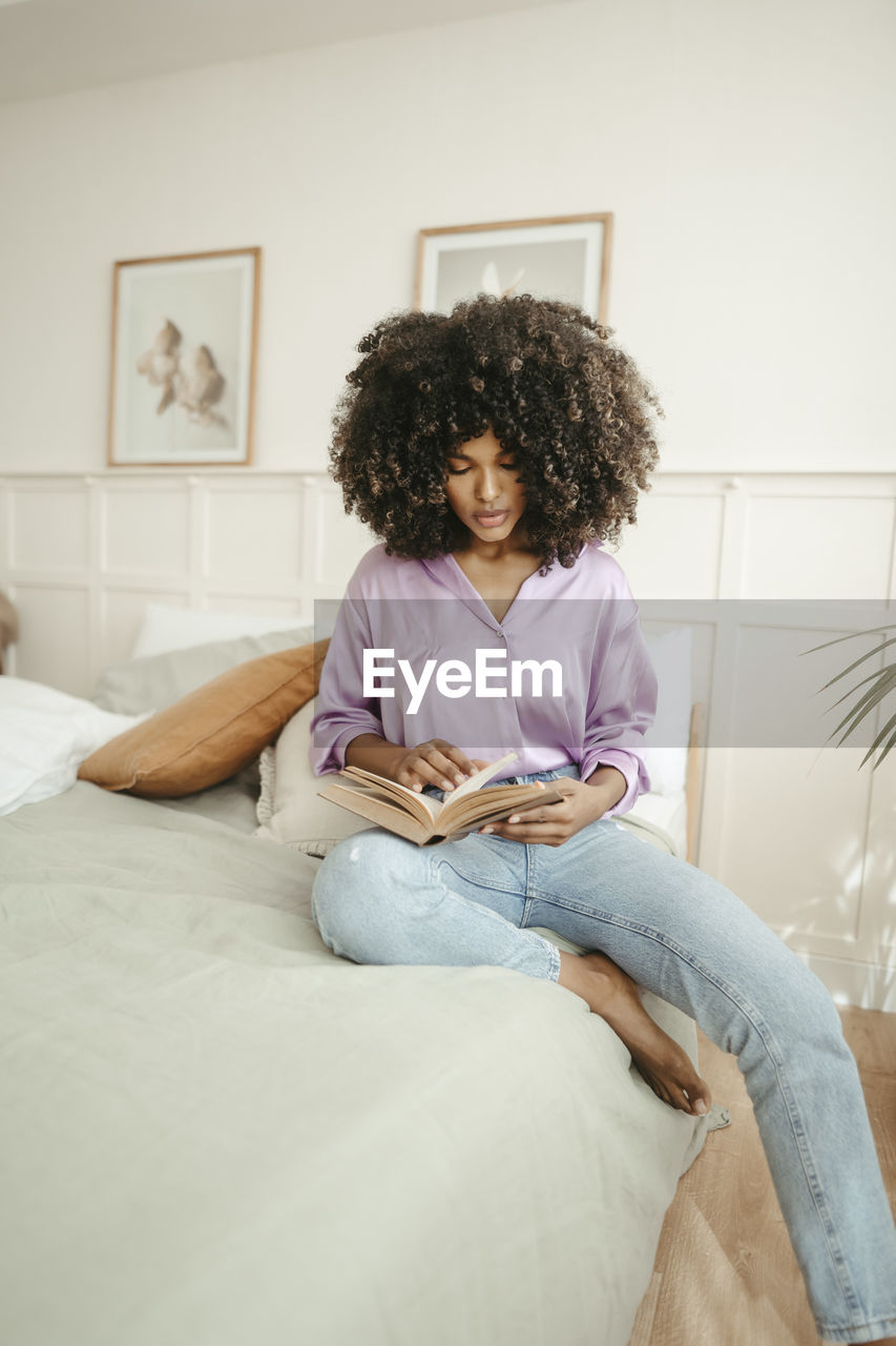 Young woman reading book sitting on bed at home