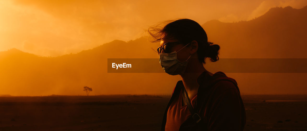 Woman wearing face mask standing against orange sky at sunset