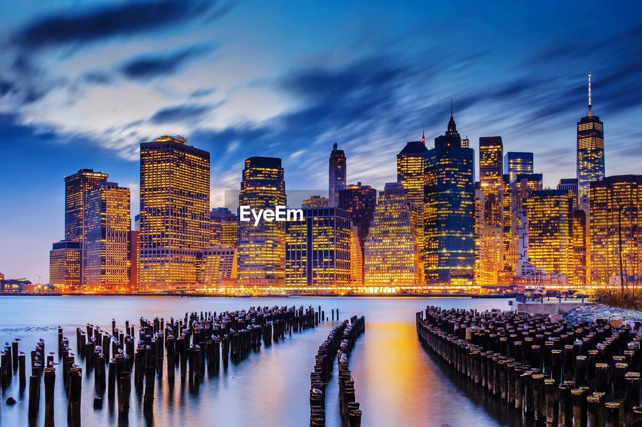 Illuminated buildings by river against sky in city at dusk