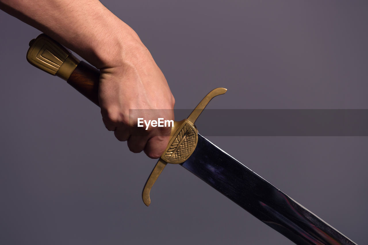 Cropped hand of man holding sword against gray background