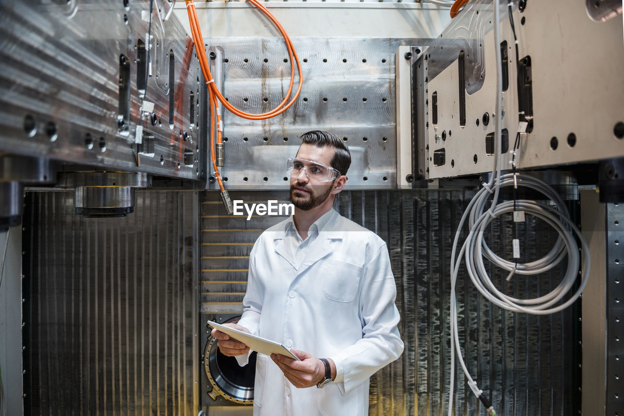 Man wearing lab coat and safety goggles at machine holding tablet