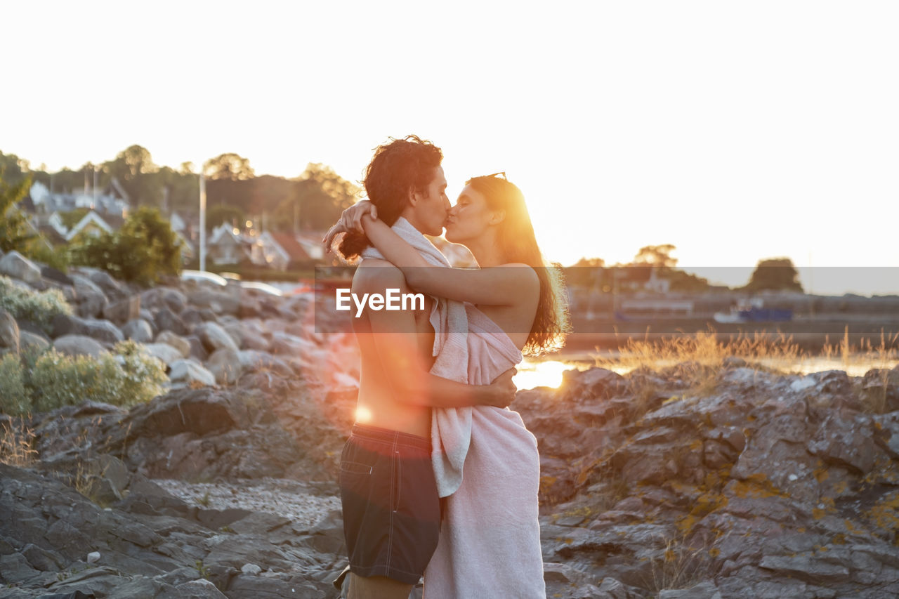 Young couple kissing while standing on rock at seashore during sunset