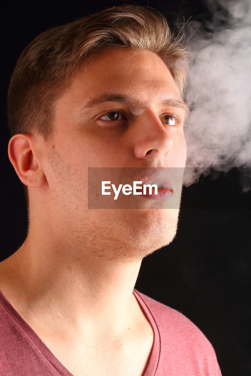 Close-up of young man exhaling smoke against black background