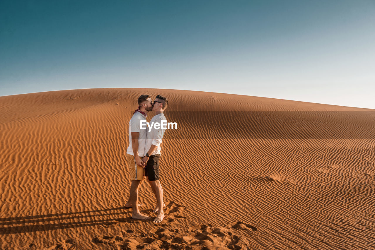 Side view of gay couple kissing while standing at desert against blue sky
