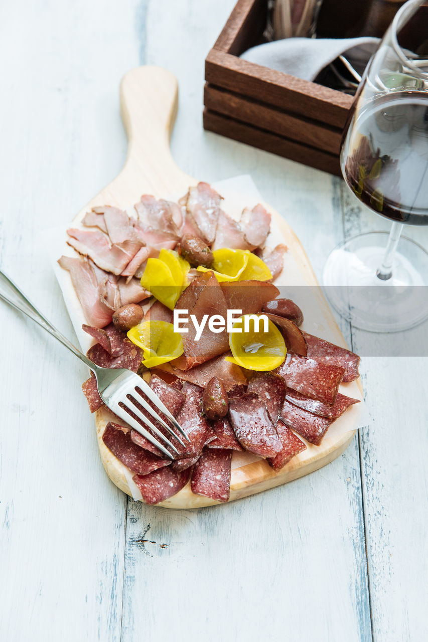 From above of slices of smoked meat different varieties creative served on wooden cutting board with metal fork and glass of red vine