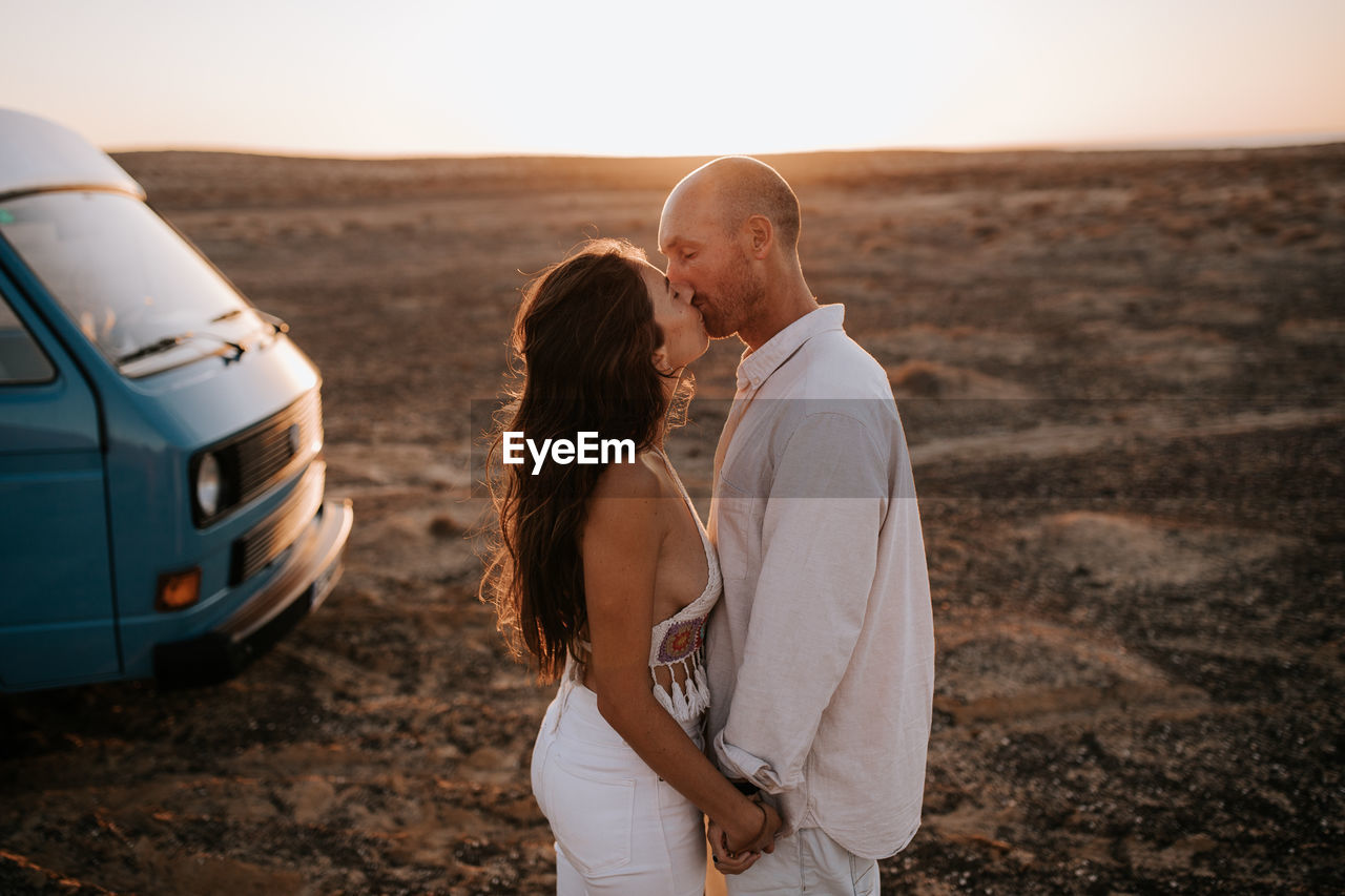 Side view of traveling couple standing near van in savanna and gently kissing on background of sunset sky